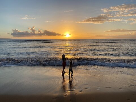 Two sisters play at the ocean&rsquo;s edge at sunset, silhouetted on a sandy beach with gentle waves, reflections, and carefree family fun.