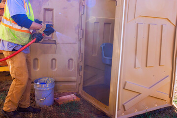 Worker sprays water inside portable restroom with pressure washer while standing on grass.