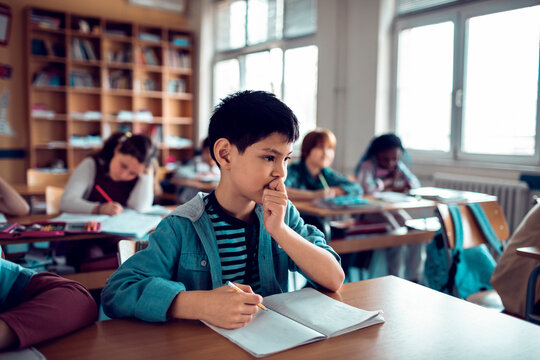 Thoughtful schoolboy writing in classroom during lesson