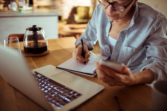 Woman taking notes while checking smartphone at kitchen table
