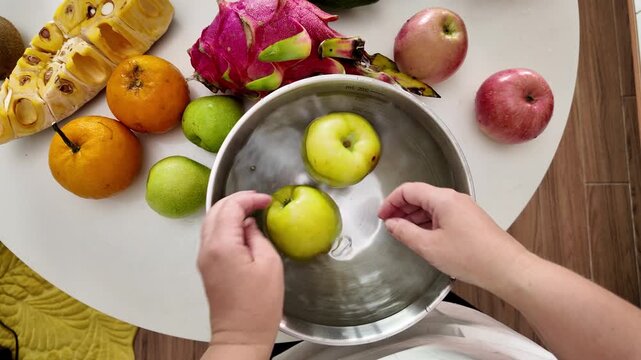Close-up of a woman's hands in an apron, standing in front of a table with fruit and washing green apples