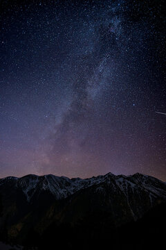 Milky Way galaxy stretching across starry night sky over dark mountain silhouette, meteor streaks visible in deep space astrophotography with purple and navy tones.