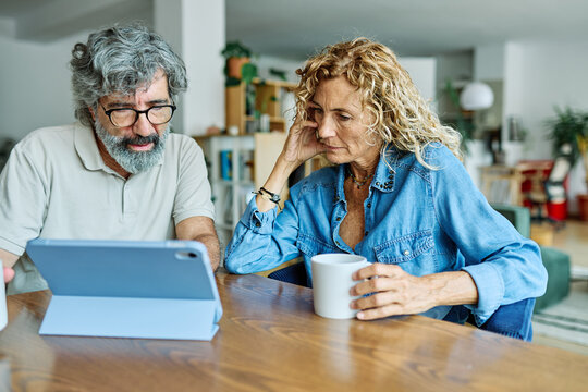 Portrait of a lovely tablet senior mature couple using a laptop together and having fun drinking coffee or tea sitting at a table at home