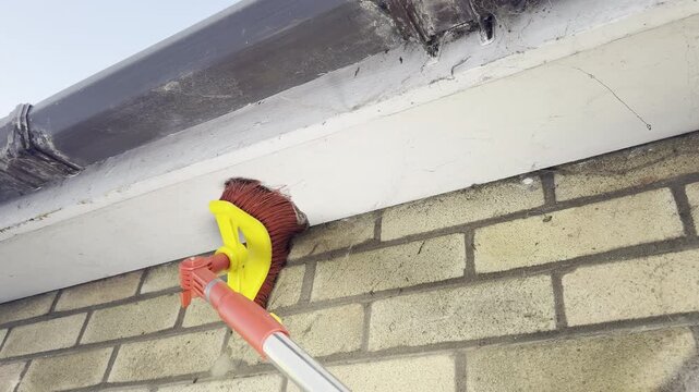 Closeup of a property maintenance job, with a special square head broom attachment, angled on a pole, sweeping along house guttering and woodwork, at the top of a brick wall, to clean cobwebs away.