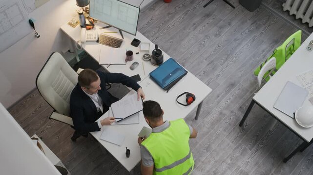 Top down shot of multiethnic adult foreman in safety vest receiving payment from male manager at table in small construction office