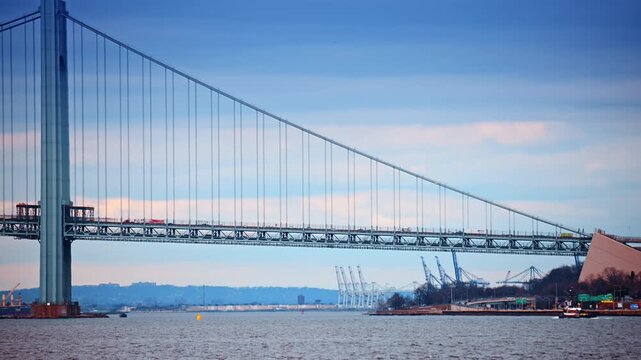 View on the Verrazzano-Narrows Bridge in New York, USA with transport moving by. Marine terminals with cranes in the backdrop.