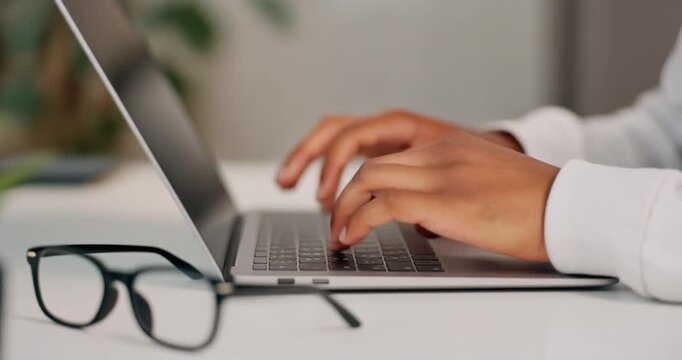 Man's hands typing on silver MacBook keyboard on white table in home office. Adult with light brown skin wearing white shirt, glasses resting on desk, green plant in background