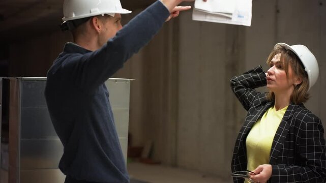 Two construction professionals, man and woman wearing hard hats, have tense argument over architectural blueprints and project details inside an unfinished concrete building
