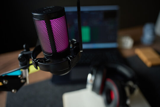 Illuminated studio microphone on boom arm on desk with headphones and laptop with audio editing timeline in background. Concept of podcast recording, streaming setup and audio content creation