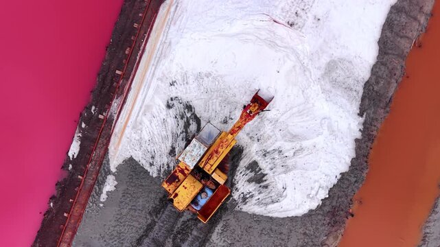 Yellow excavator digging into a large mound of white salt next to pink water. Directly above aerial view of excavator harvesting salt.