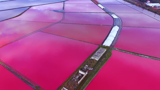 Wide drone shot of multiple pink evaporation ponds with old concrete structures and white salt ridges. Aerial landscape of salt pans and abandoned industrial buildings.