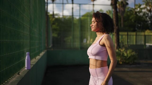Athletic woman stretching leg against fence on outdoor tennis court, focused posture during warm down, sunlight and greenery emphasizing flexibility, recovery and fitness routine.