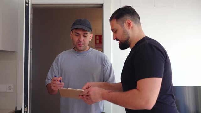 Home service specialist reviews notes on clipboard while talking to male customer near doorway. Indoor residential setting emphasizes repair coordination and maintenance planning.