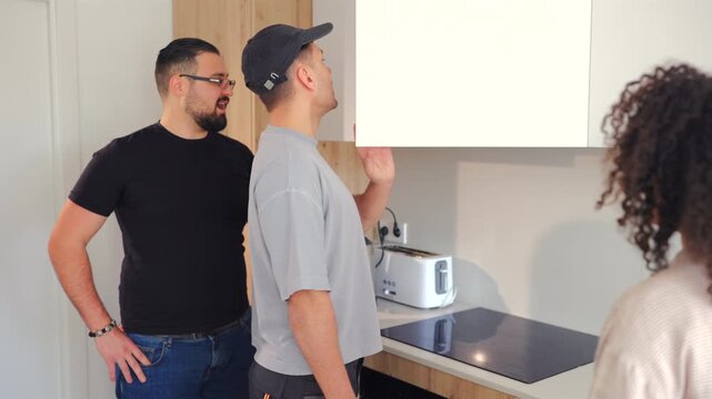 Handyman with toolbox talks to homeowners about kitchen cabinet alignment and installation process during renovation consultation inside contemporary apartment interior.