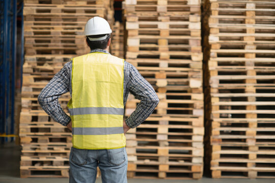 Warehouse worker wea hard hat and safety vest looking at a stack of wooden pallets in a large distribution warehouse area ready for shipping.