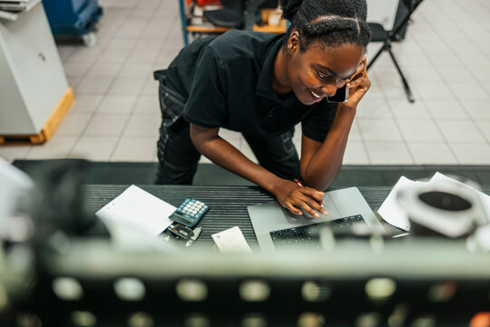 High angle view of smiling young female worker talking on smart phone while using laptop in factory plant