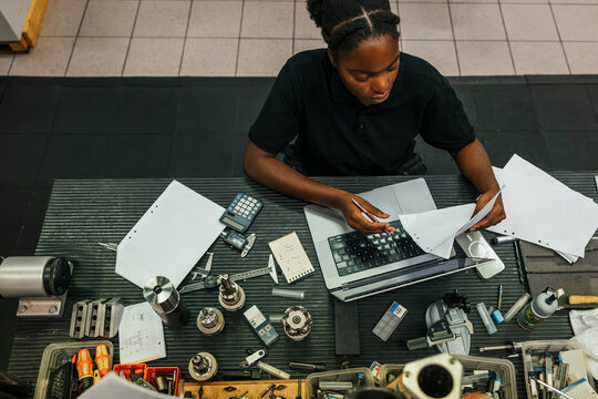 High angle view of focused young female worker reading documents while using laptop at manufacturing industry
