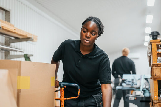 Focused female worker pushing hand truck of cardboard boxes in factory