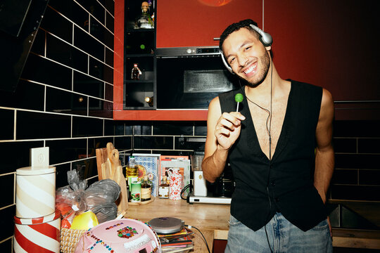 Portrait of smiling man listening to music while holding lollipop in kitchen at home