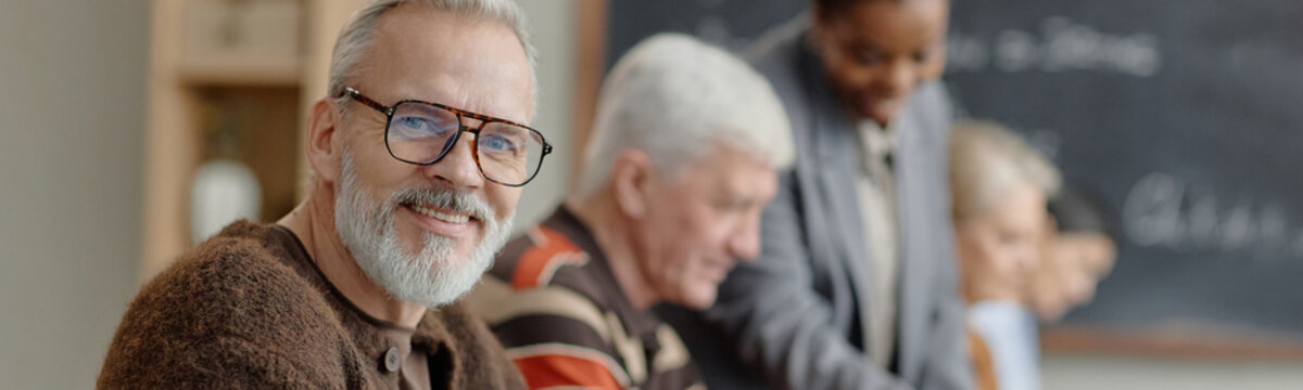 Website header shot of smiling senior Caucasian man looking at camera while sitting in classroom with group of senior adults studying together, Black woman assisting at chalkboard