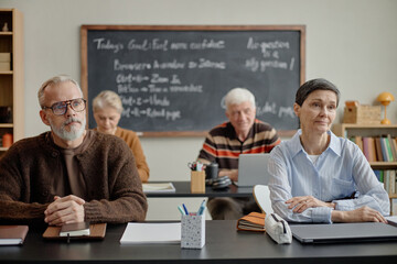 Group of senior Caucasian men and women attending class, sitting at desks and listening...