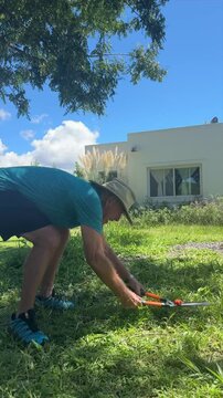Elderly man gardening cutting grass with hedge shears in sunny yard
