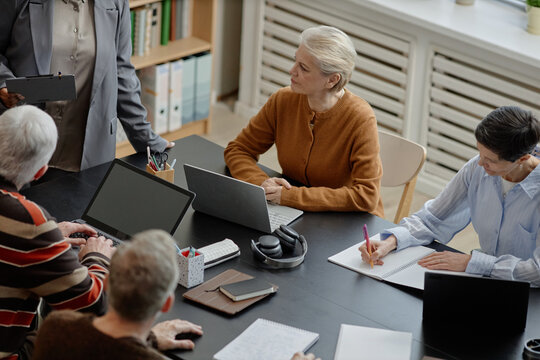 Group of middle aged and senior Caucasian women and men participating in educational meeting, sitting at table with laptops and notebooks, listening and taking notes during discussion