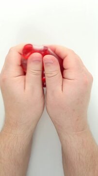 Two hands stretch out and knead the red squish sad, anti-stress, stretching it wide and then squeezing it into a rounded mass on a white background. Elastic resistance and wet pops provide ASMR fun.