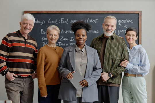 Group of smiling middle aged and senior adults posing next to Black female teacher standing together in classroom with blackboard