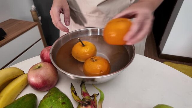 Hands of a woman in an apron standing in front of a table with fruit and washing mandarins