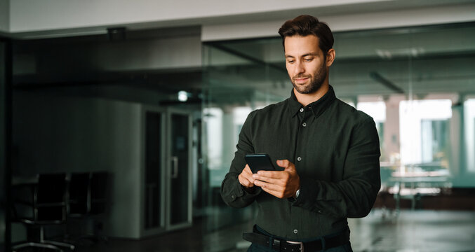 Young businessman holding smartphone telephone device standing in office. European ceo manager trader using digital technology cellphone phone mobile for business trade work. Web banner, copy space