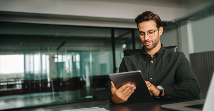 European man working on trade online using touchpad fintech computer for business work at workplace. Young entrepreneur businessman holding digital pc tablet sitting in office. Web banner, copy space