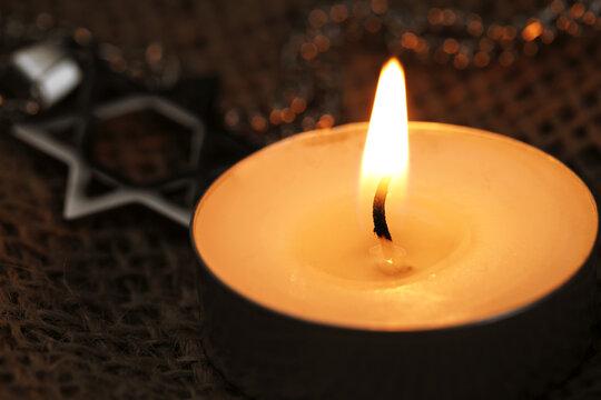 Holocaust Remembrance Day. Burning candle and pendant in shape of Star of David on burlap cloth, selective focus