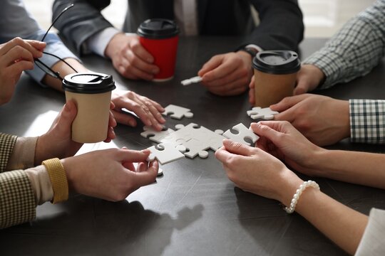 Team building. Group of businesspeople with takeaway cups solving puzzle together at dark wooden table indoors, closeup