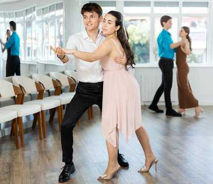 Expressive Hispanic guy in formal attire practicing sensual bachata with attractive young brunette in light flowing dress in dance class. Social dancing concept..