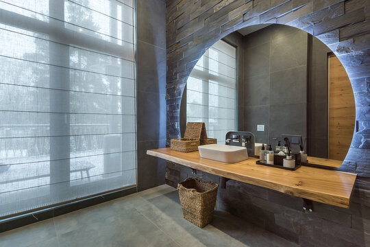 A bathroom showcases a round mirror above a wooden counter with a sink. Baskets hold toiletries, and large windows let in natural light. The space has a clean and simple feel