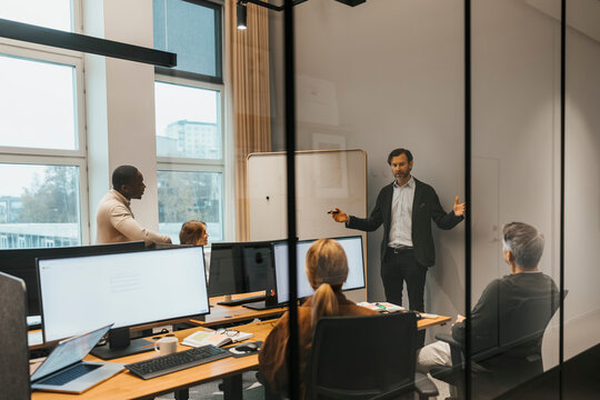 Male entrepreneur explaining business strategies to team in meeting room seen through glass