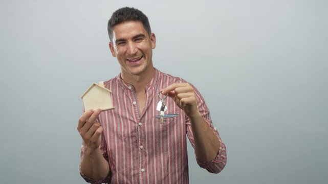 Young hispanic man smiling and holding a small wooden house and a set of keys in a pale blue studio; homeownership joy.