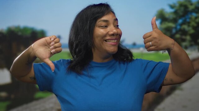 Woman wearing blue shirt shows thumbs up and thumbs down gesture in sunlit park with blurred trees; mixed conflict duality ambivalence.