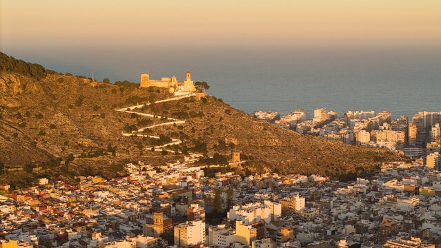 Aerial drone view of Cullera in Spain during golden hour, featuring a hilltop castle and winding path above the Mediterranean Sea. Detailed coastal cityscape with rooftops, shoreline and dramatic suns