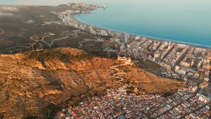 Fototapeta premium Aerial drone view of Cullera in Spain during golden hour, featuring a hilltop castle and winding path above the Mediterranean Sea. Detailed coastal cityscape with rooftops, shoreline and dramatic suns
