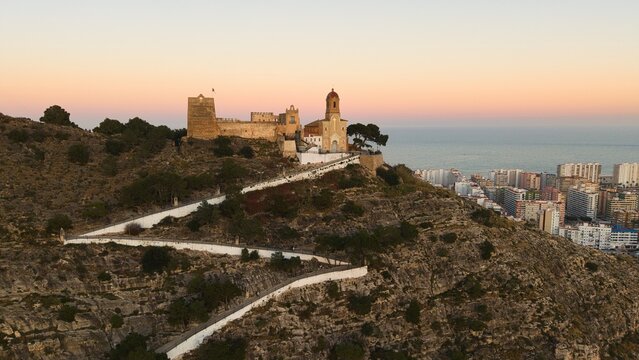 Aerial drone view of Cullera in Spain during golden hour, featuring a hilltop castle and winding path above the Mediterranean Sea. Detailed coastal cityscape with rooftops, shoreline and dramatic suns
