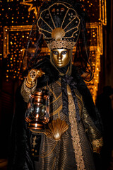 Venice, Italy - February 11, 2026: Elegant masked figure in ornate black and gold costume holding lantern at Venice Carnival illuminated by festive lights