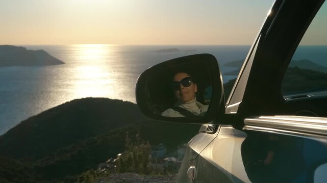 Woman's reflection in car mirror at sunset overlooking the sea. Close up of a woman's face reflected in a car's side-view mirror, enjoying a beautiful scenic drive along a coastal road with the sun