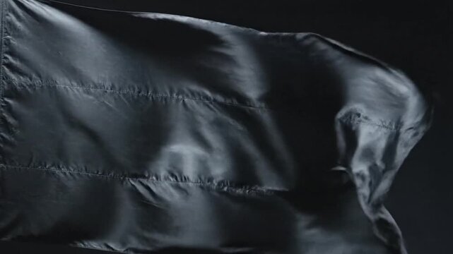 A plain black flag made of fabric material waving gently in the wind against a dark background.