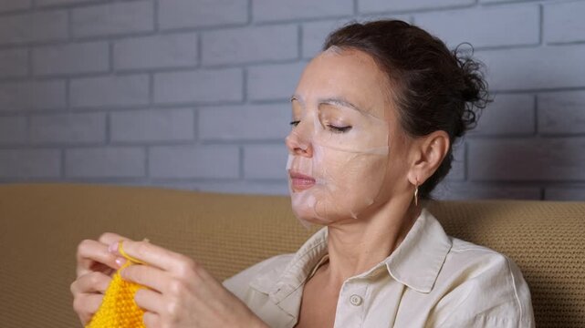 Woman relaxing with face mask knitting at home. Relaxed woman with a cosmetic sheet mask on her face knitting with yellow yarn while sitting on the sofa at home