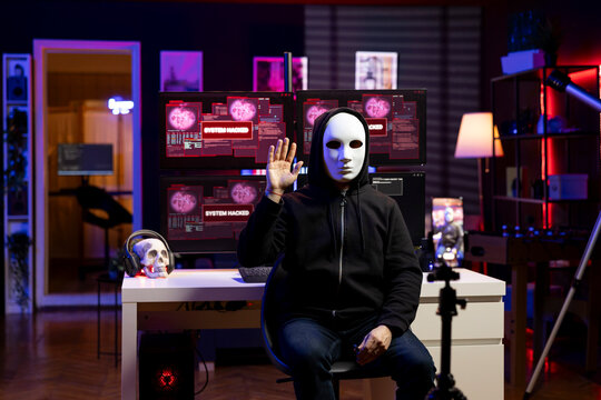 Masked hacker greeting audience, filming ransom video in hidden home. Cybercriminal concealing identity using phone on tripod to record blackmail footage in neon lit hideout