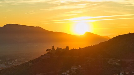 Obraz premium Cinematic aerial view of Cullera in Spain at sunset with castle and church silhouetted against golden light. Mediterranean coastline and mountain backdrop create a dramatic coastal cityscape.
