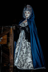 Obraz premium Venice, Italy - February 11, 2026: Woman in ornate masquerade costume with blue cloak and intricate dress stands by wooden railing during Venice Carnival night celebration