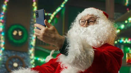 Upbeat Santa Claus doing videocall with kids using smartphone, bringing joy. Santa Claus...